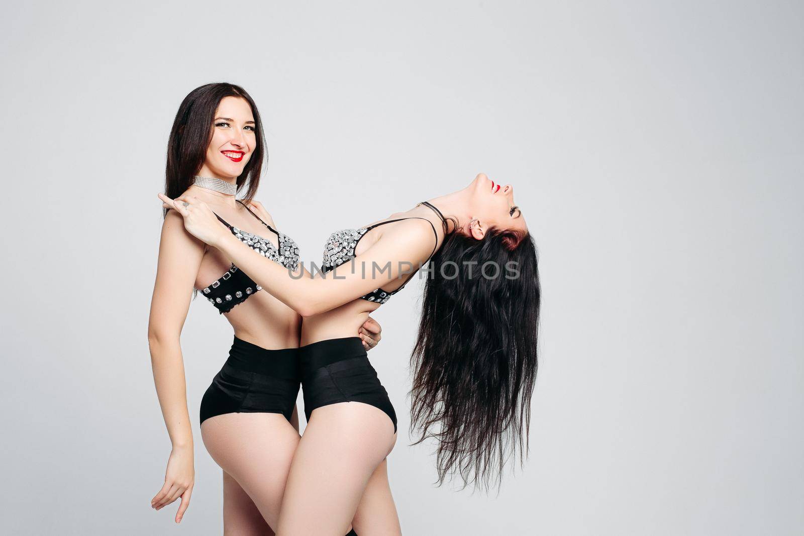 Studio portrait of two happy seductive girls with long black hair wearing sexy dancing costumes hugging over white background.
