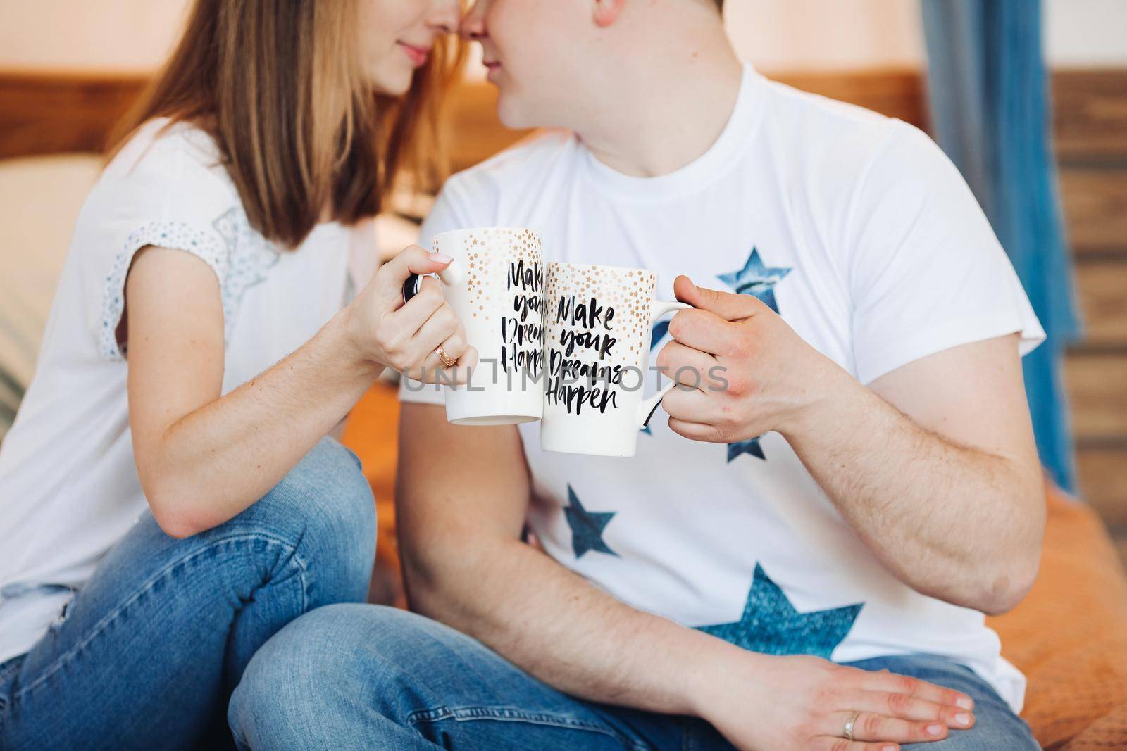 Portrait of romantic couple in love, sitting on bed decorated with lights, with creative cup in hand. Boyfriend and girlfriend wearing in white t shirt and jeans embracing and looking at camera.