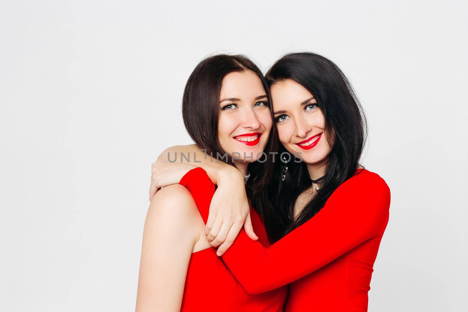 Beautiful smiling sisters in red embracing over white background. by StudioLucky