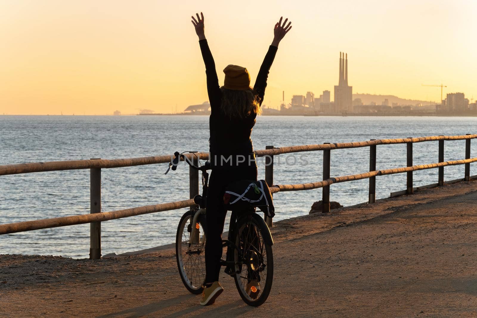 Happy girl with a bicycle raised her hands up and enjoys the sunset on the promenade road of Maresme, Catalonia, Spain.