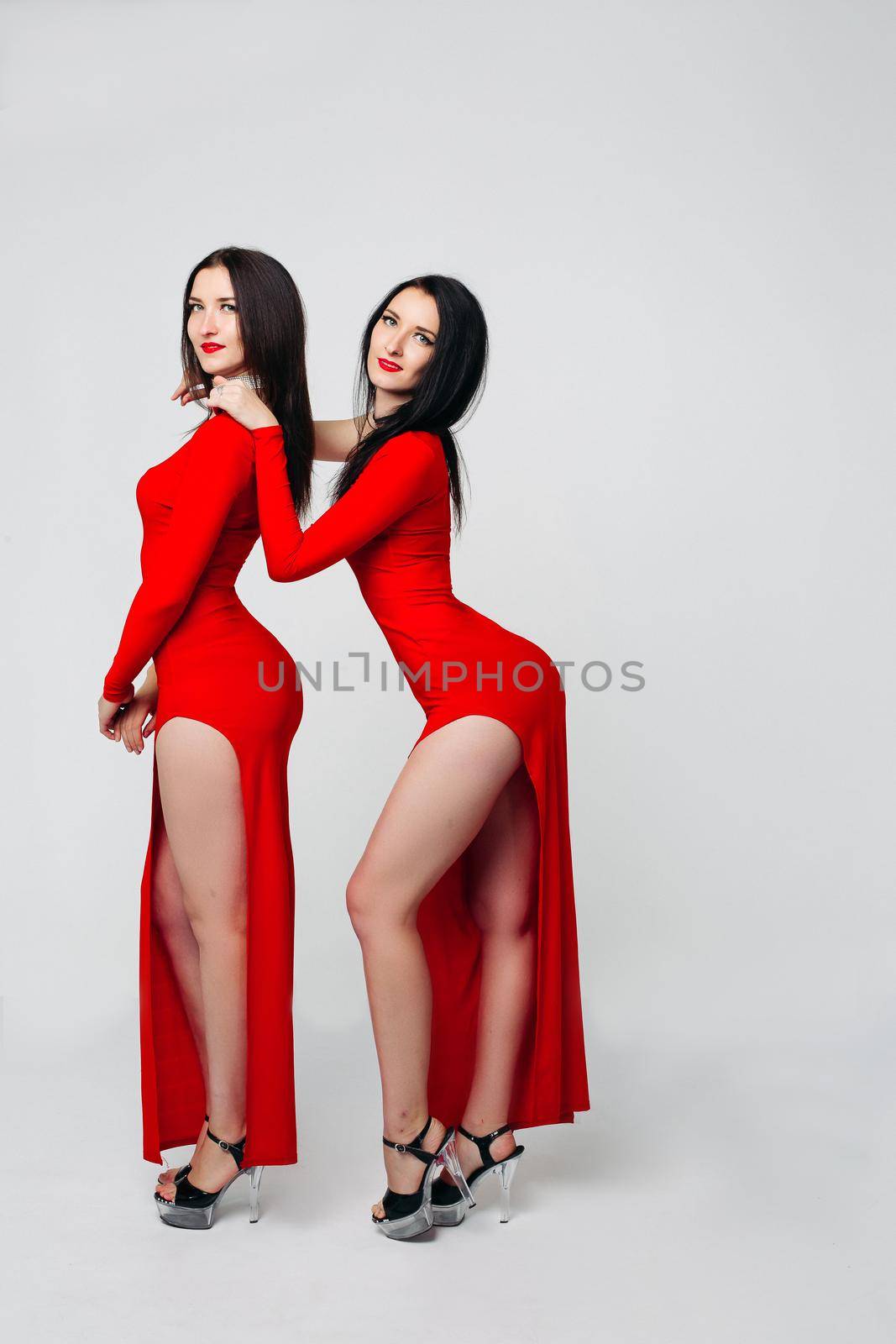 Studio full length of two sexy twins with black hair and bright make-up wearing long red dresses and high heels for pole dancing over white background. Looking at camera.