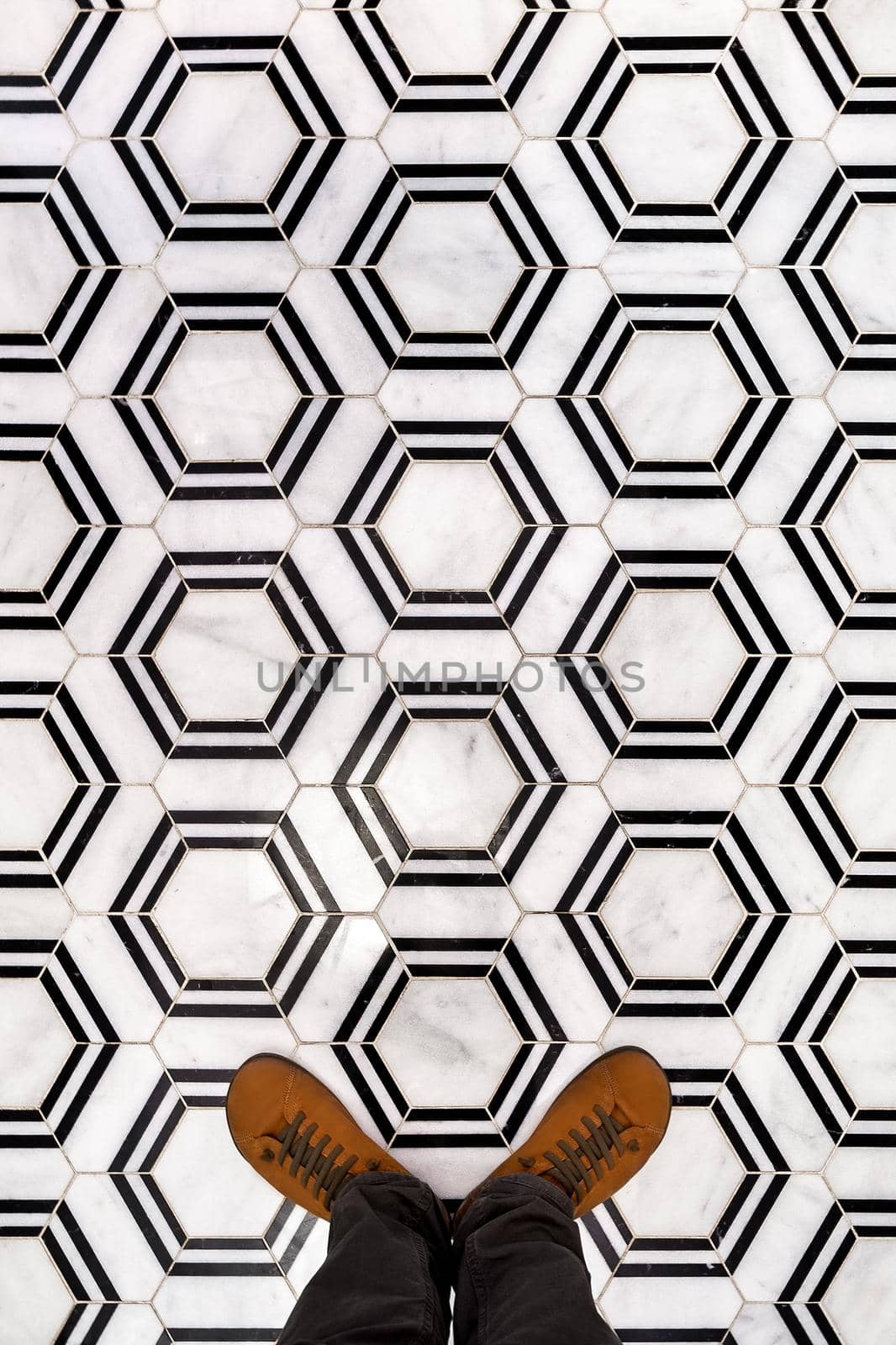 Top view of men's legs in black jeans wear brown leather shoes, standing on black and white hexagon floor tiles.
