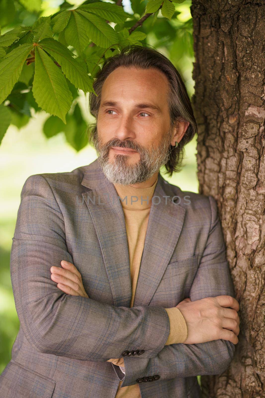 Handsome mature grey birded man in casual standing under tree on the grass looking away with arms folded. Freelancer spend time working outdoors.