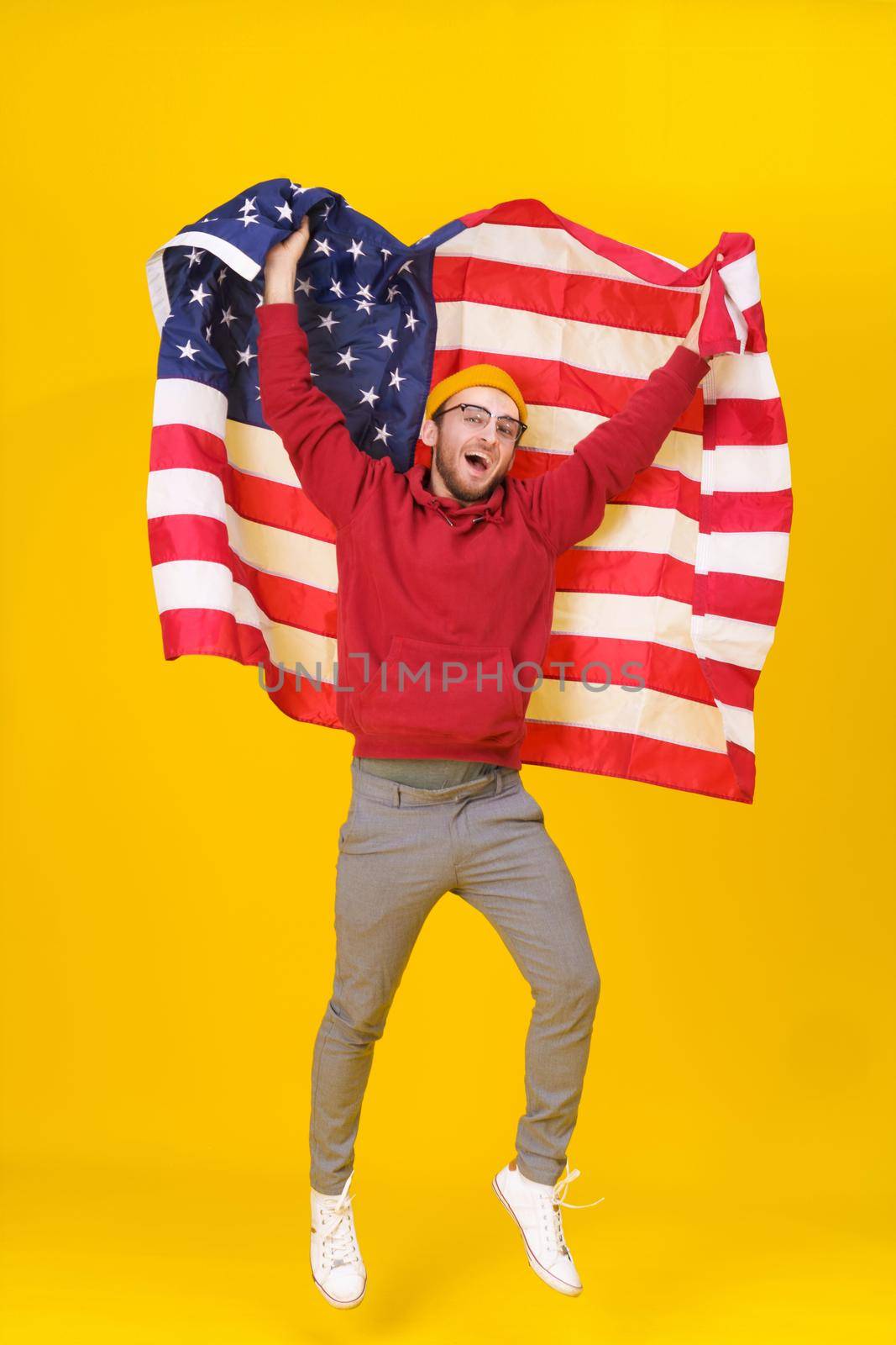Cheerful young man with American flag. Happy, funny young man in red hoodie and USA flag jumping hight isolated on yellow background. freedom is in your life by LipikStockMedia