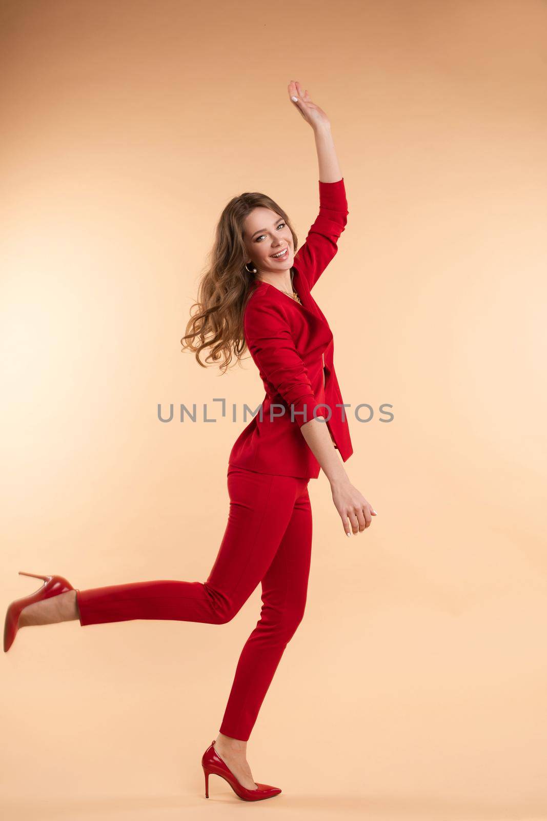 Side view of happy young woman in red smart suit looking at camera and jumping in studio. Beautidul lady on heels laughing and posing on isolated background. Concept of beauty and happiness.
