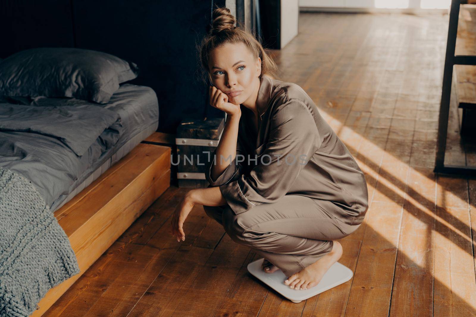Image from up of young upset woman measuring her weight, dressed in satin pajama squatted on electronic scales in early morning after waking up in bedroom. Weight Loss and dieting concept