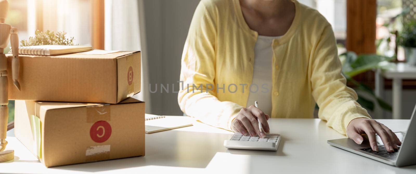 Close up asian female entrepreneur, checks online orders and uses a price calculator to calculate product prices and packs products into boxes to customers