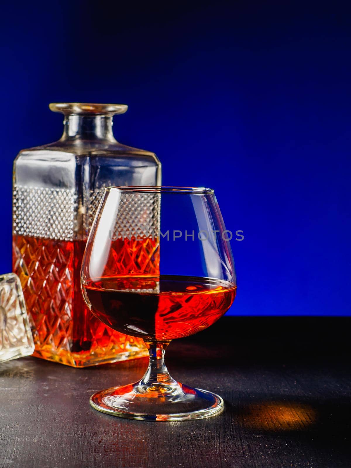 glass textured square bottle and glass goblet with amber alcohol brandy scotch whiskey on a dark blue glowing background. Vertical photography