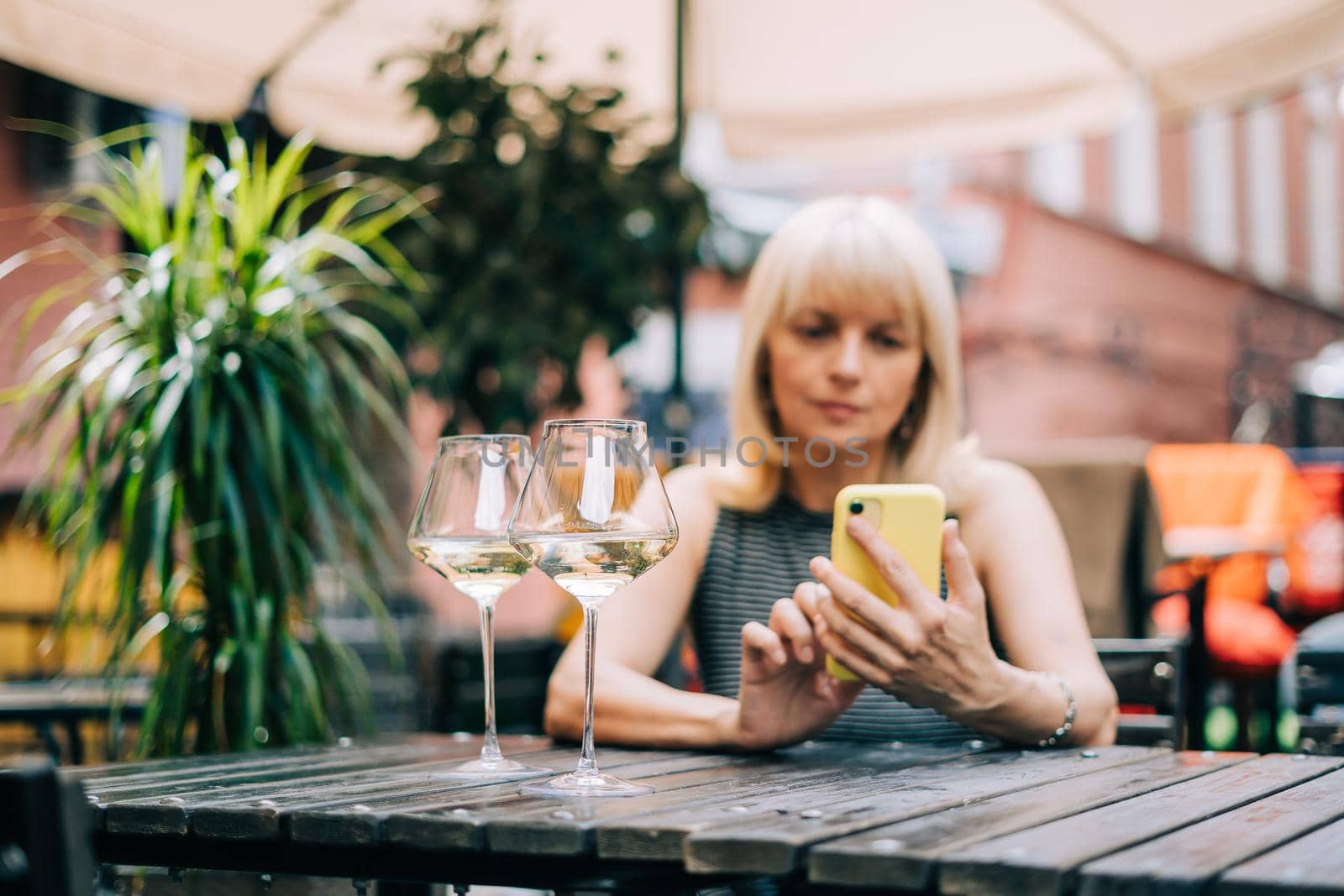 Adult mature woman texting messaging at mobile phone in bar outdoors with wine glasses and blurry restaurant, drinking white wine. People using technology cellphone. Lifestyle at summer sunny day