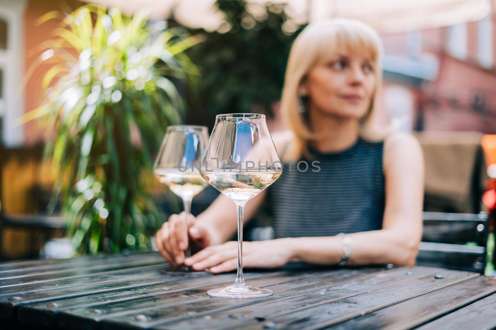 Thoughtful adult mature woman sitting in bar outdoors with wine glasses and blurry restaurant background scene, drinking white wine. Summer sunny day on patio. People lifestyle