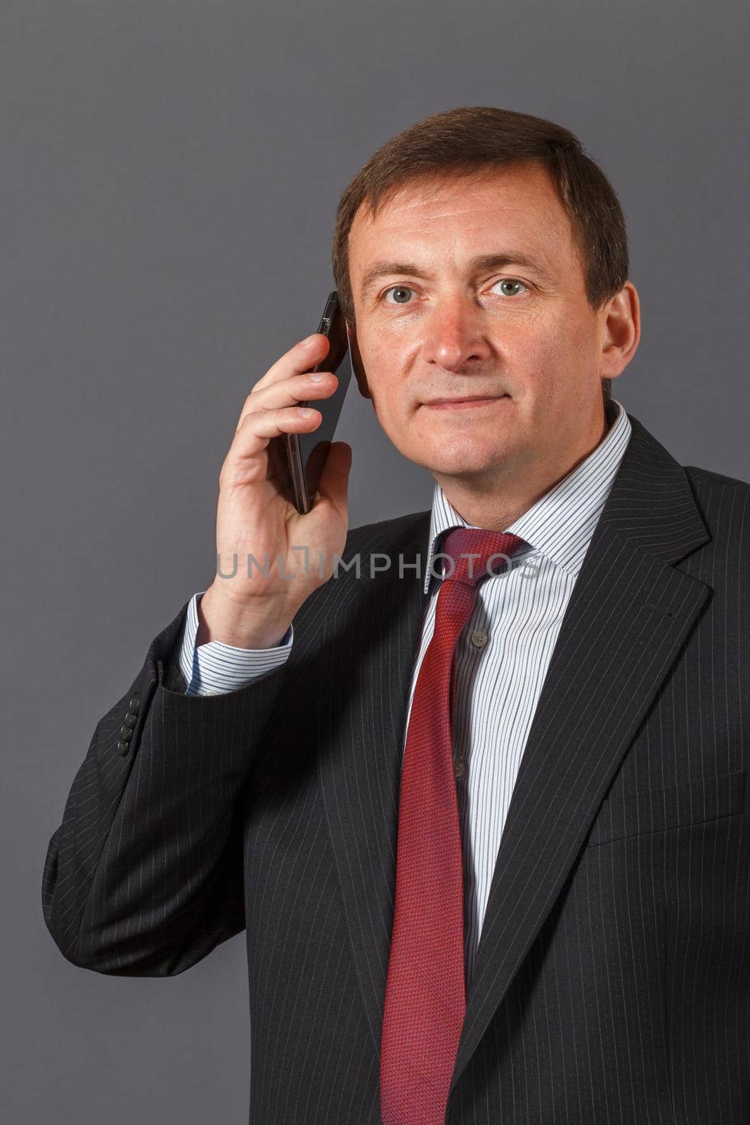Confident elegant handsome mature businessman standing in front of a gray background in a studio wearing a nice suit is speaking on the sell phone