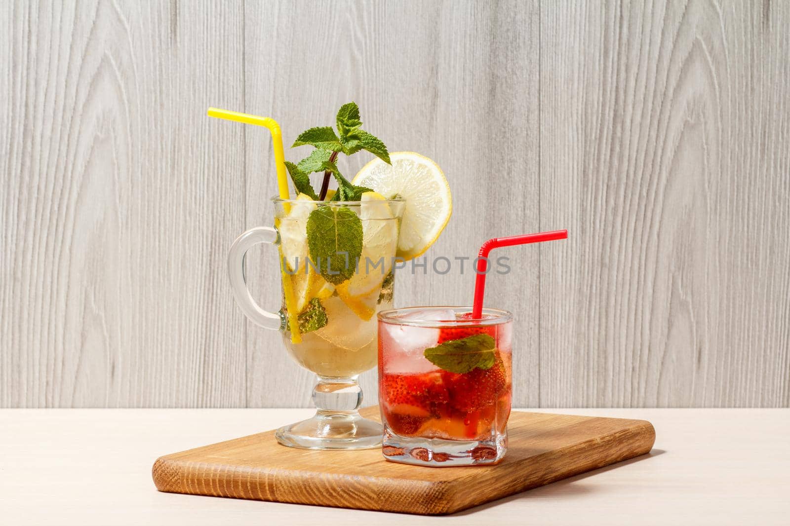 Cold refreshing summer lemonade mojito with lemon slices and mint in a tall glass and strawberry lemonade with mint in a short one on a wooden cutting board
