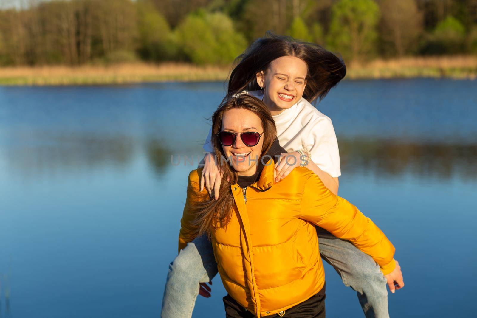 Playful mother giving daughter piggy back ride at spring lake shore. Both laughing and look happy. Spring in lake background. Closeup.