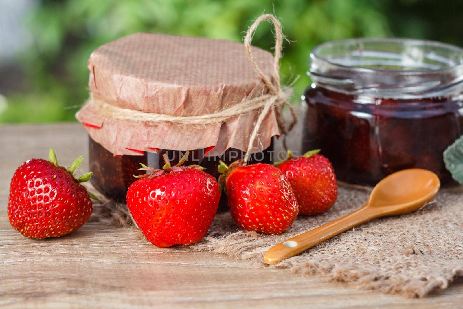 Traditional homemade strawberry jam in jars with spoon, decorated with fresh strawberries on wooden table with green blurred natural background Close up selective focus