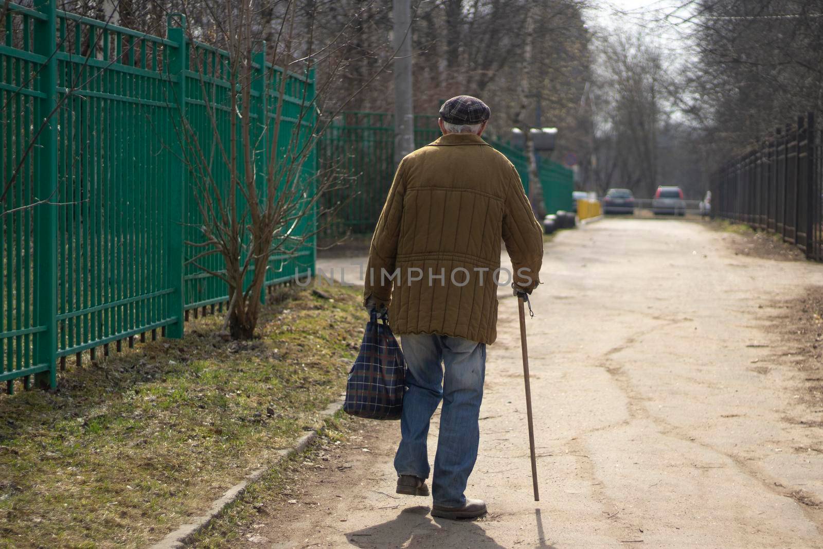 Pensioner in Russia. Poverty in world. Elderly man walks leaning on walking stick. Background man goes into distance. Old jacket and cap.