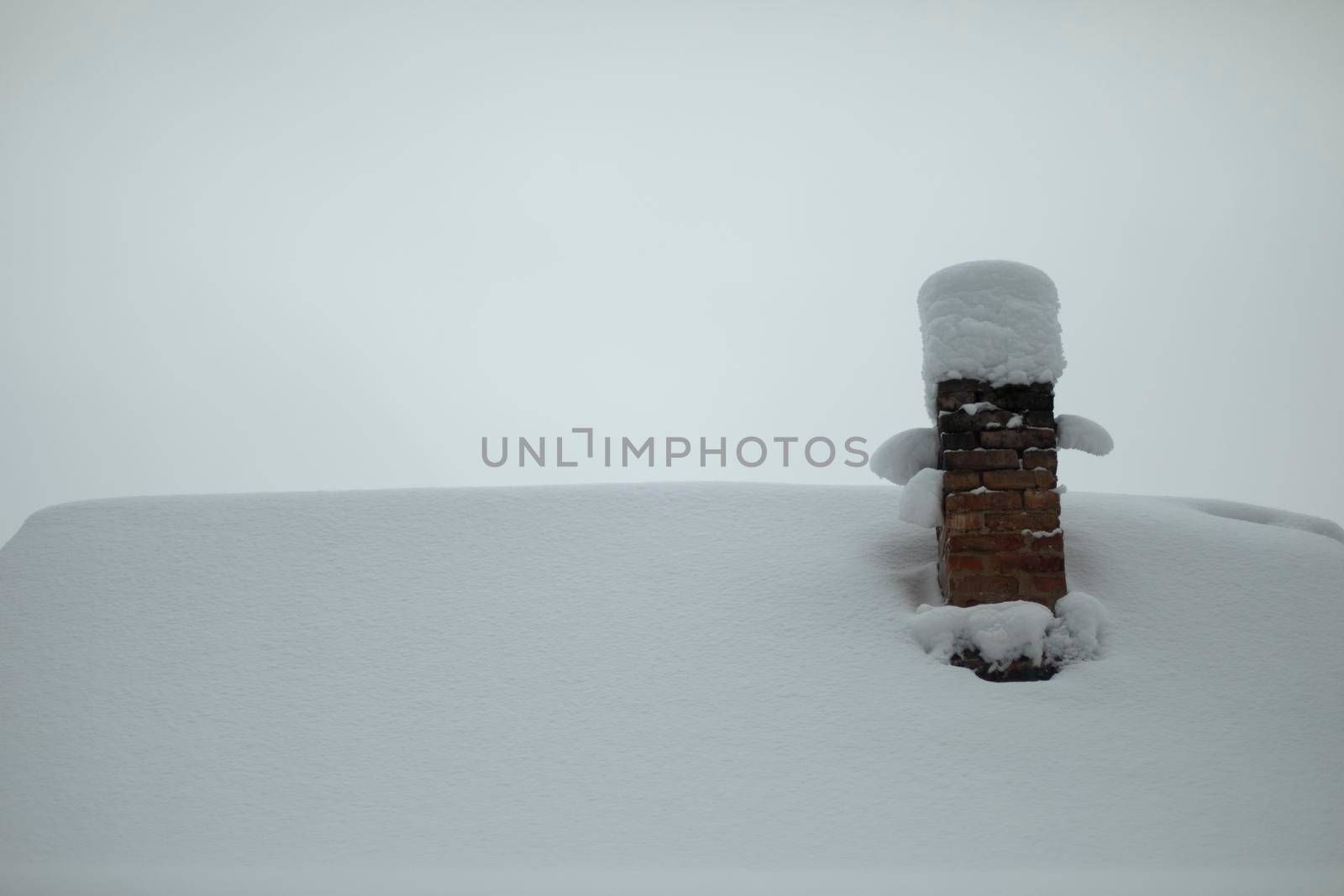 Roof is in snow and chimney. Pipe from stove in old house. Area is rural in winter. Cozy winter.