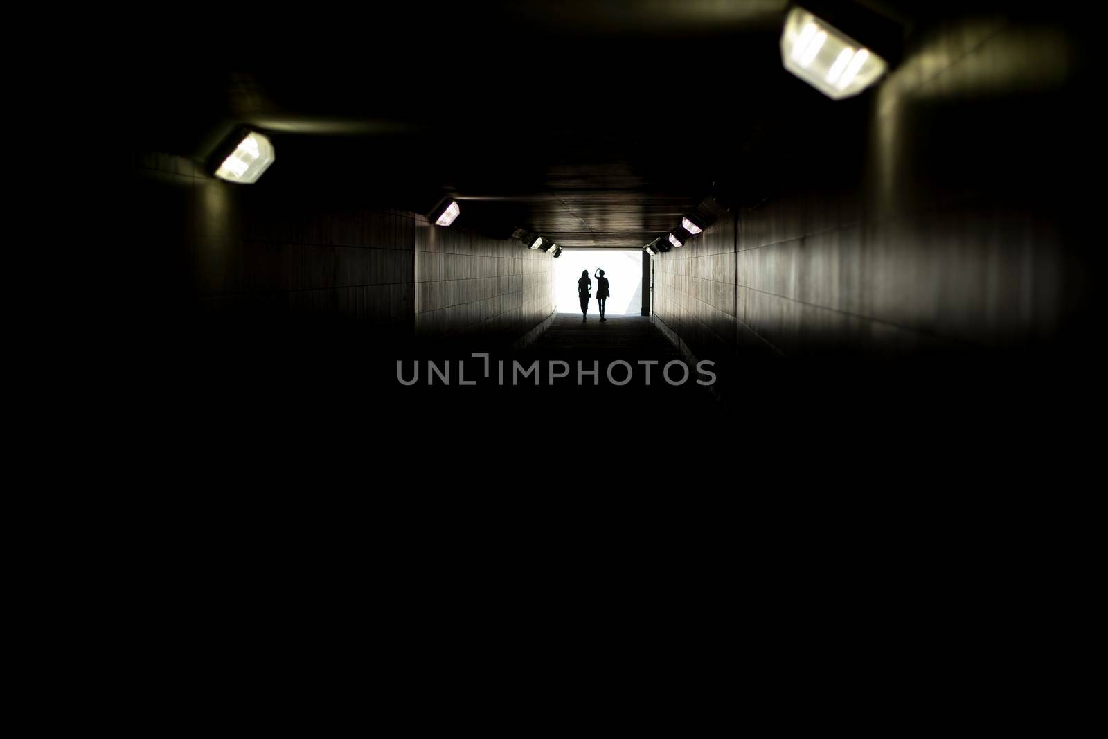 Two silhouettes at end of tunnel. Light in distance. Figures of small women in contrasting light. Lamps in long underpass.