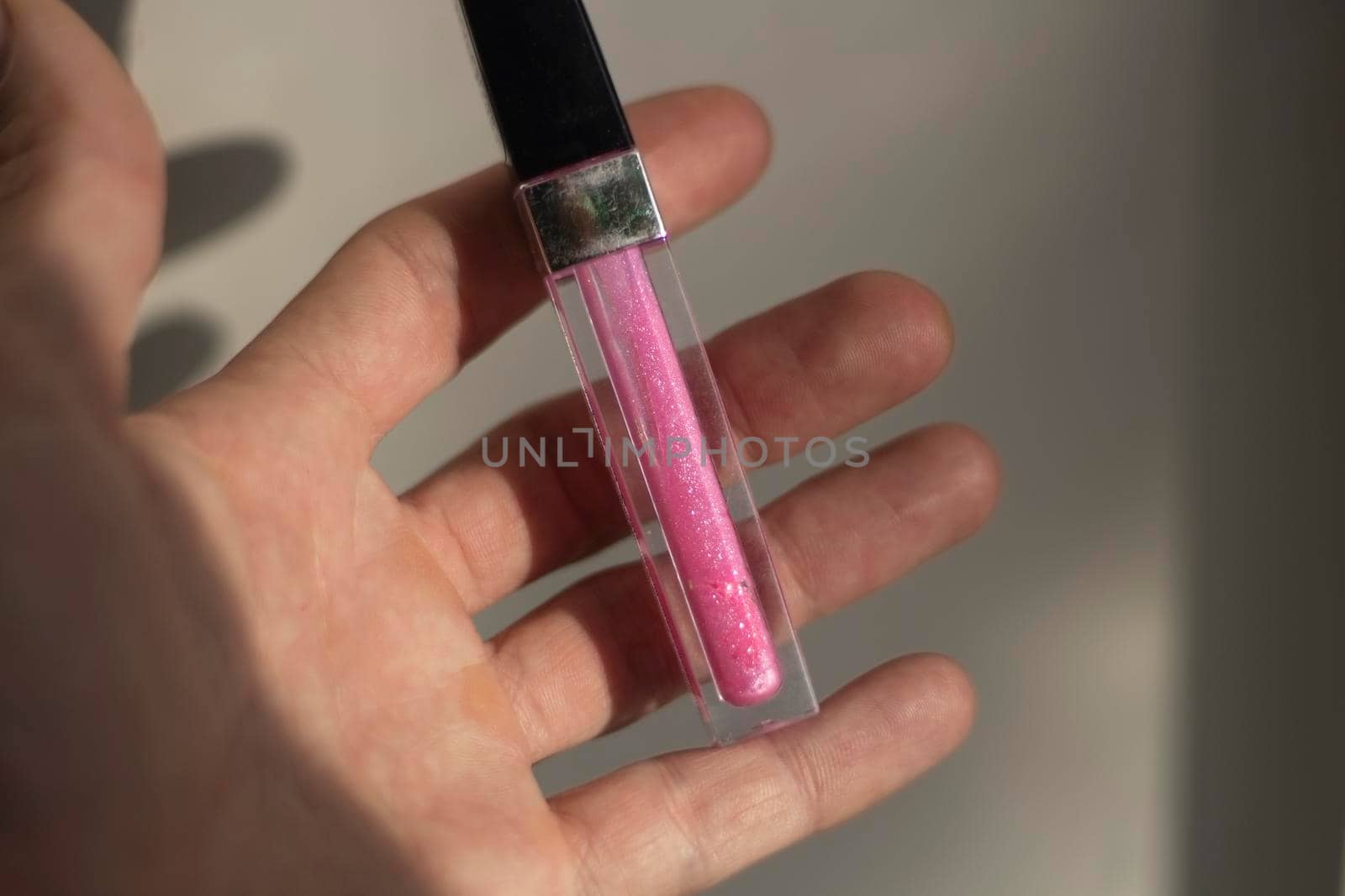 A bottle with a pink lip gloss lies in the palm of a person's hand, illuminated by sunlight. Makeup tool. Indoor shooting