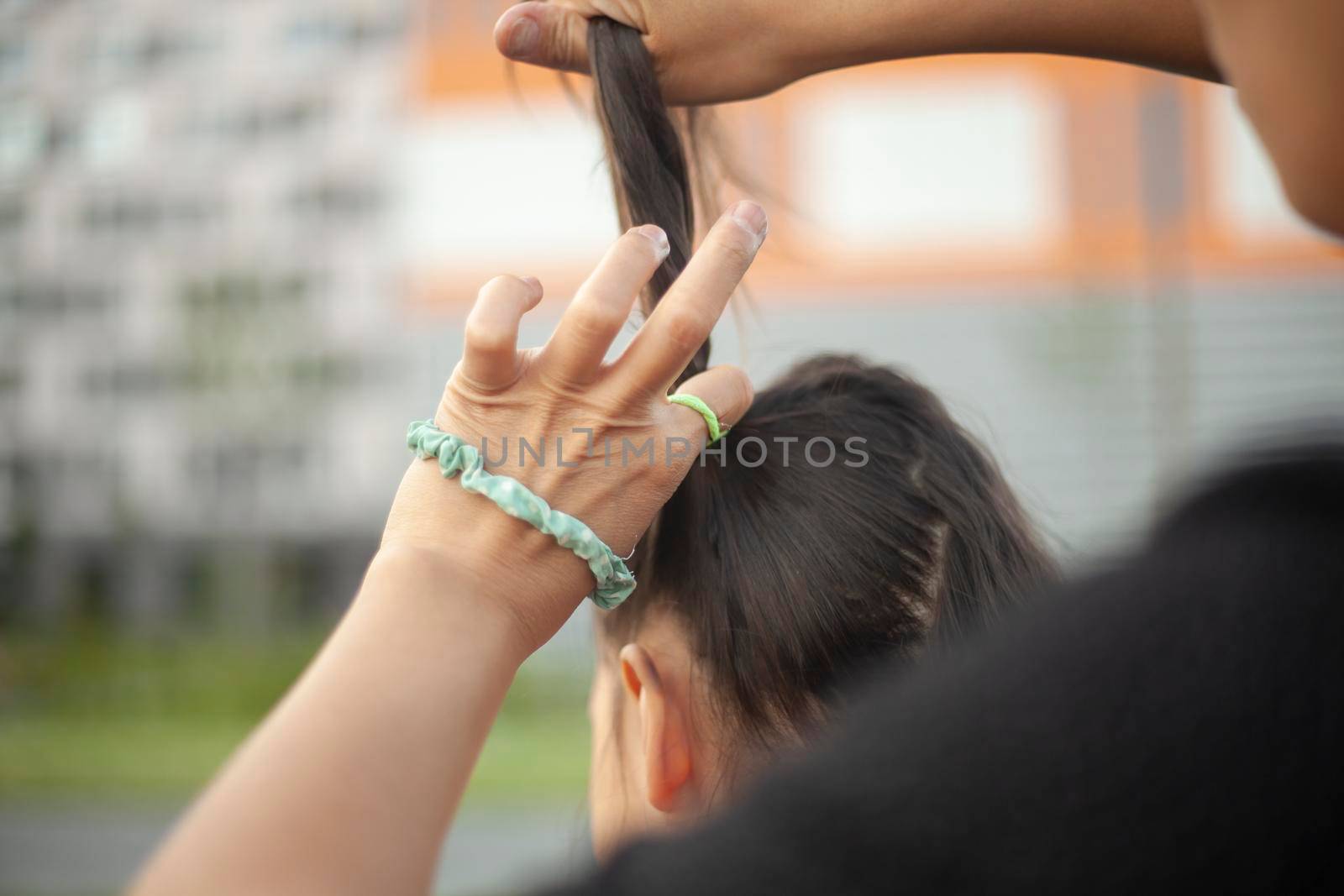 Mother makes ponytail out of her daughter's hair. Creating hairstyle for child. Elastic band for thick hair. Care for preschooler. Help with hair styling. Child on street.
