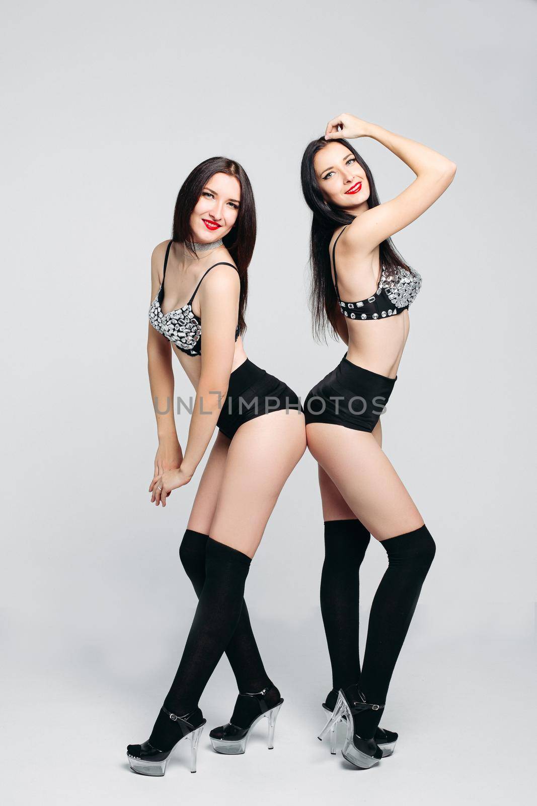 Studio portrait of beautiful sexy twins posing in seductive pole dance or striptease costumes and high heels, smiling at camera.