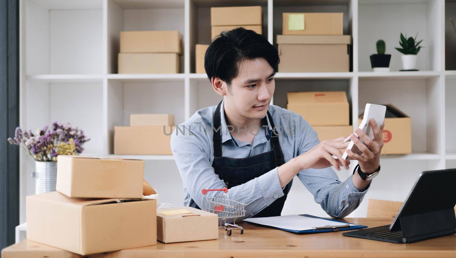 A portrait of Asian man, e-commerce employee sitting in the office full of packages on the table using a calculator, for SME business, e-commerce, technology and delivery business..