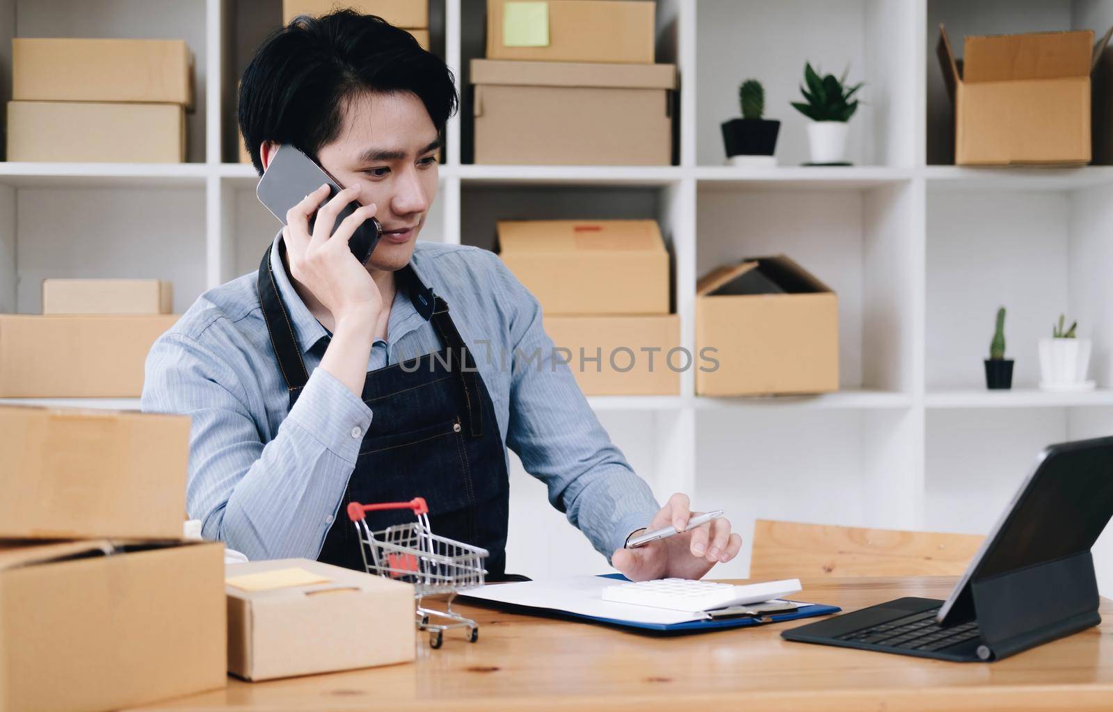 Young asia seller man happy work on laptop busy cellphone call chat to customer at home office desk use digital tablet computer in SME online store social e-commerce sale page service web support. by wichayada