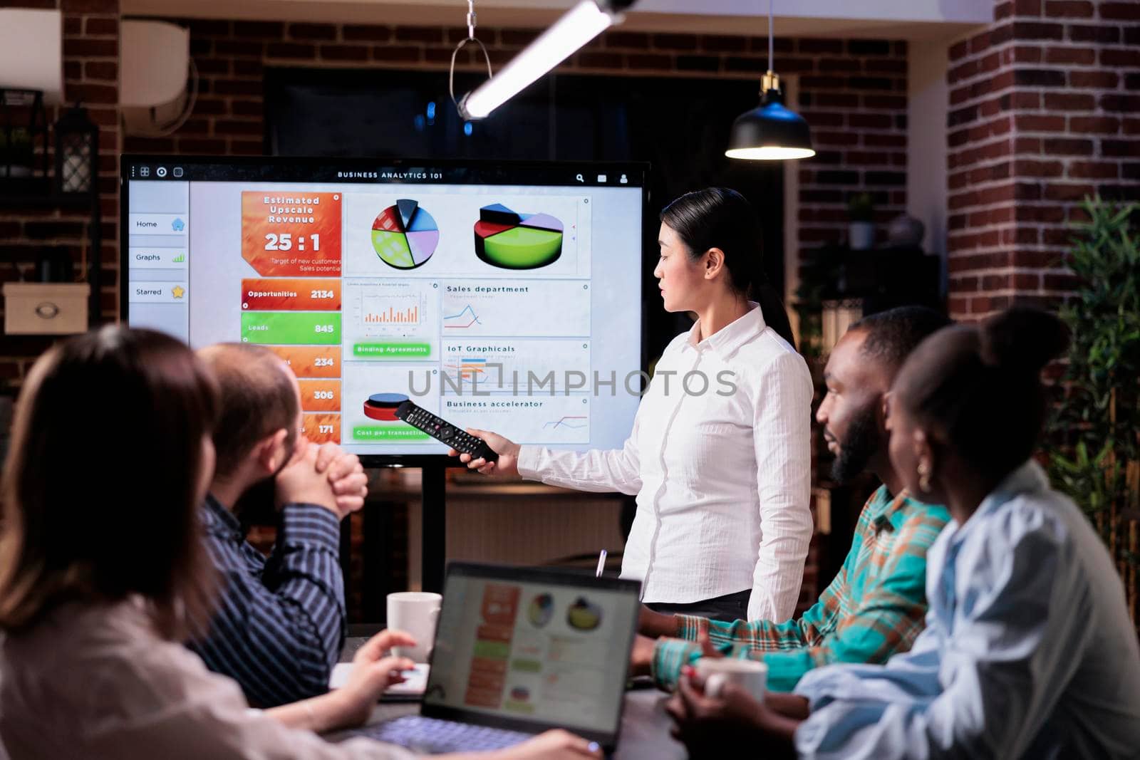 Asian entrepreneur holding remote control presenting business sales statistics on tv screen in late night meeting. Woman workahollic doing overtime at work talking about marketing strategy with diverse team.