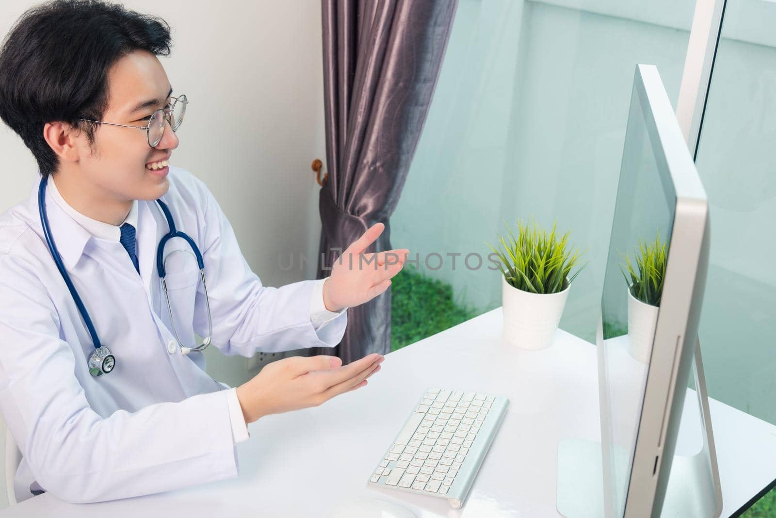 Asian young handsome doctor man wearing a doctor's dress and stethoscope video conference call or facetime raise hand to explain the symptoms he smiling at hospital office, Health medical care concept