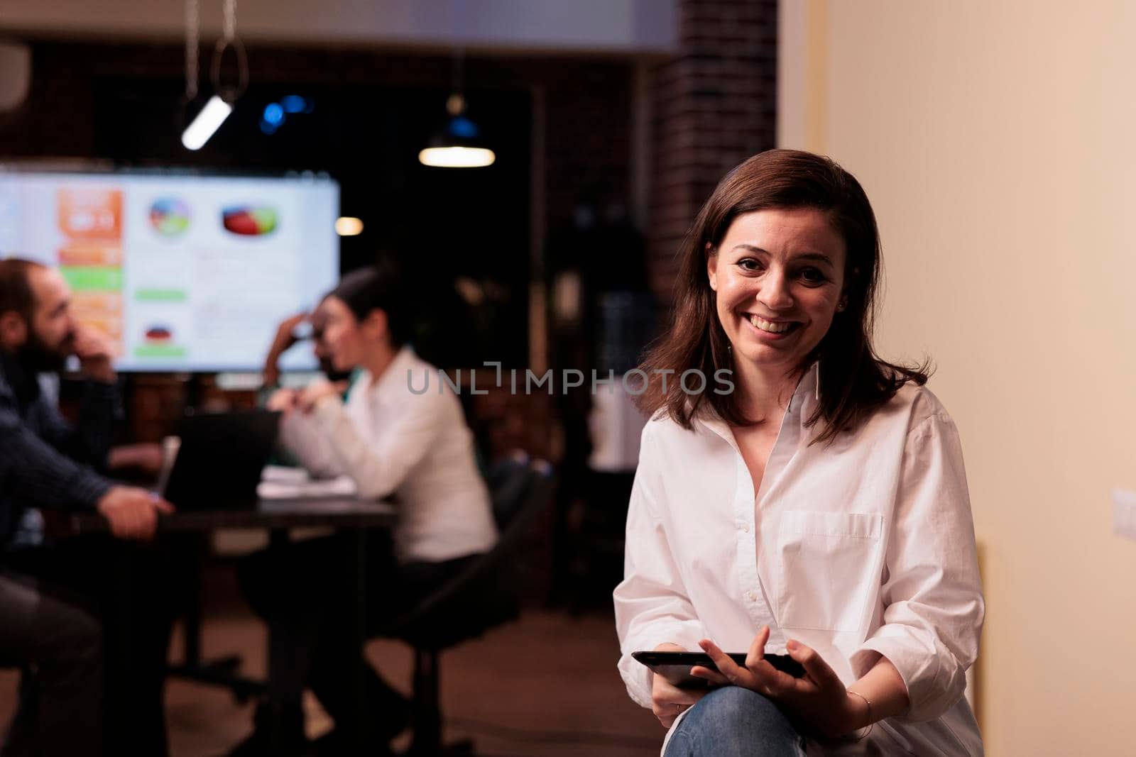 Portrait of smiling caucasian woman holding digital tablet working overtime in business office. Startup employee posing confident in busy workspace working with touchscreen device with team.