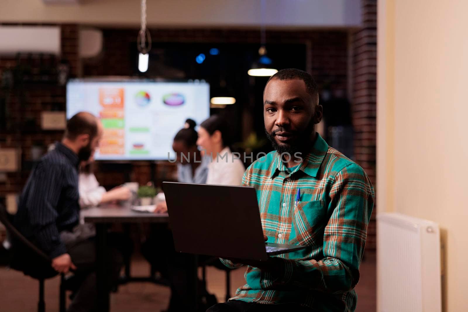 Portrait of african american man holding laptop in startup office during late night meeting in sales conference. Employee posing confident working overtime with mixed team brainstorming sales strategy.