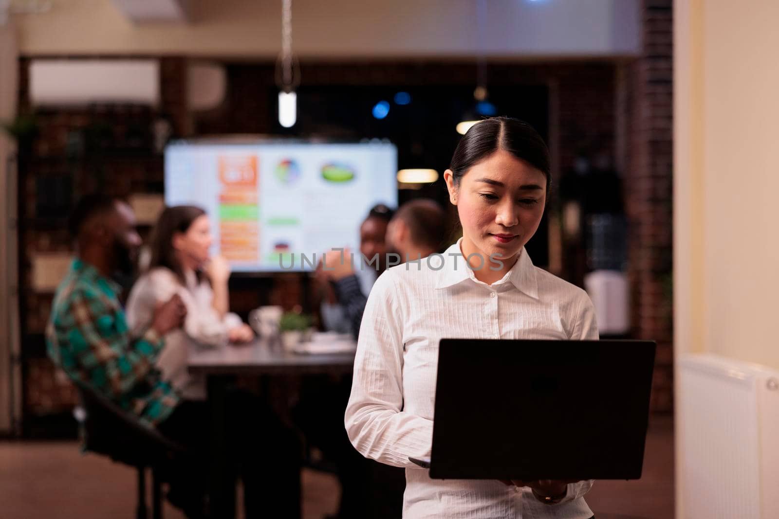 Focused asian woman holding laptop and looking at screen working late hours during business startegy meeting with mixed team. Startup employee posing confident in group project with coworkers.