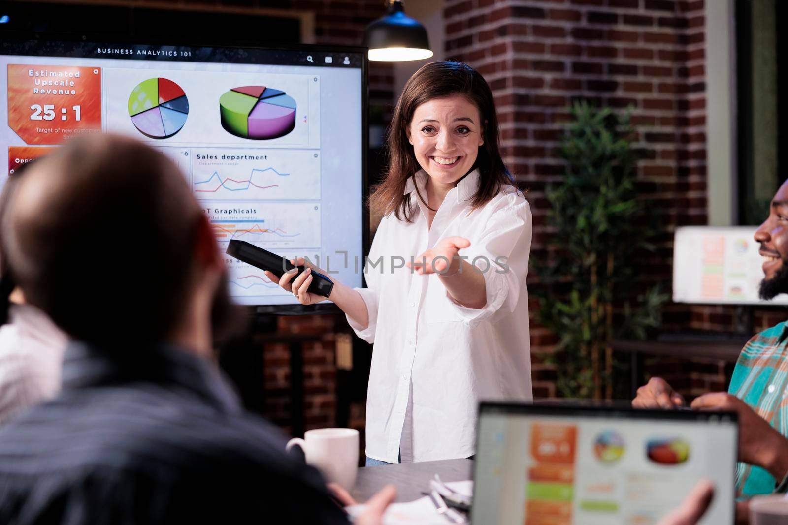 Smiling business employee standing in front of tv screen with sales statistics talking with man with laptop by DCStudio