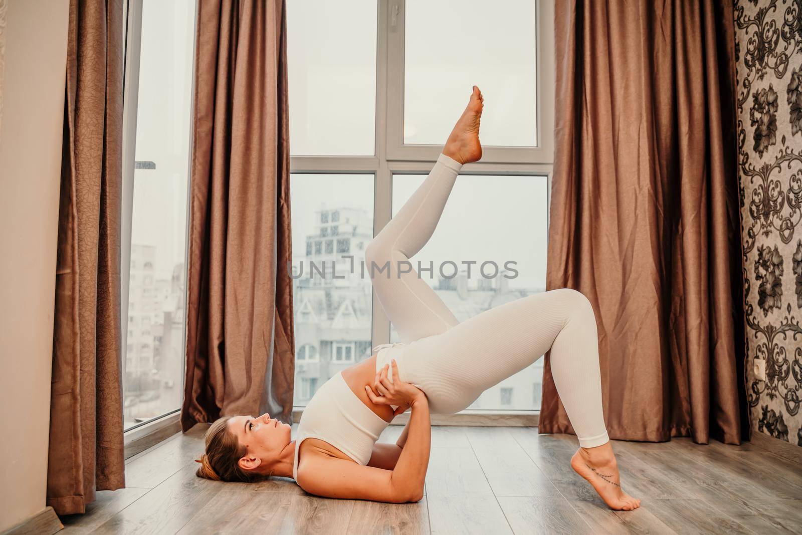 Young athletic attractive woman practicing yoga. Works out at home or in a yoga studio, sportswear, white pants and a full-length top indoors. Healthy lifestyle concept.