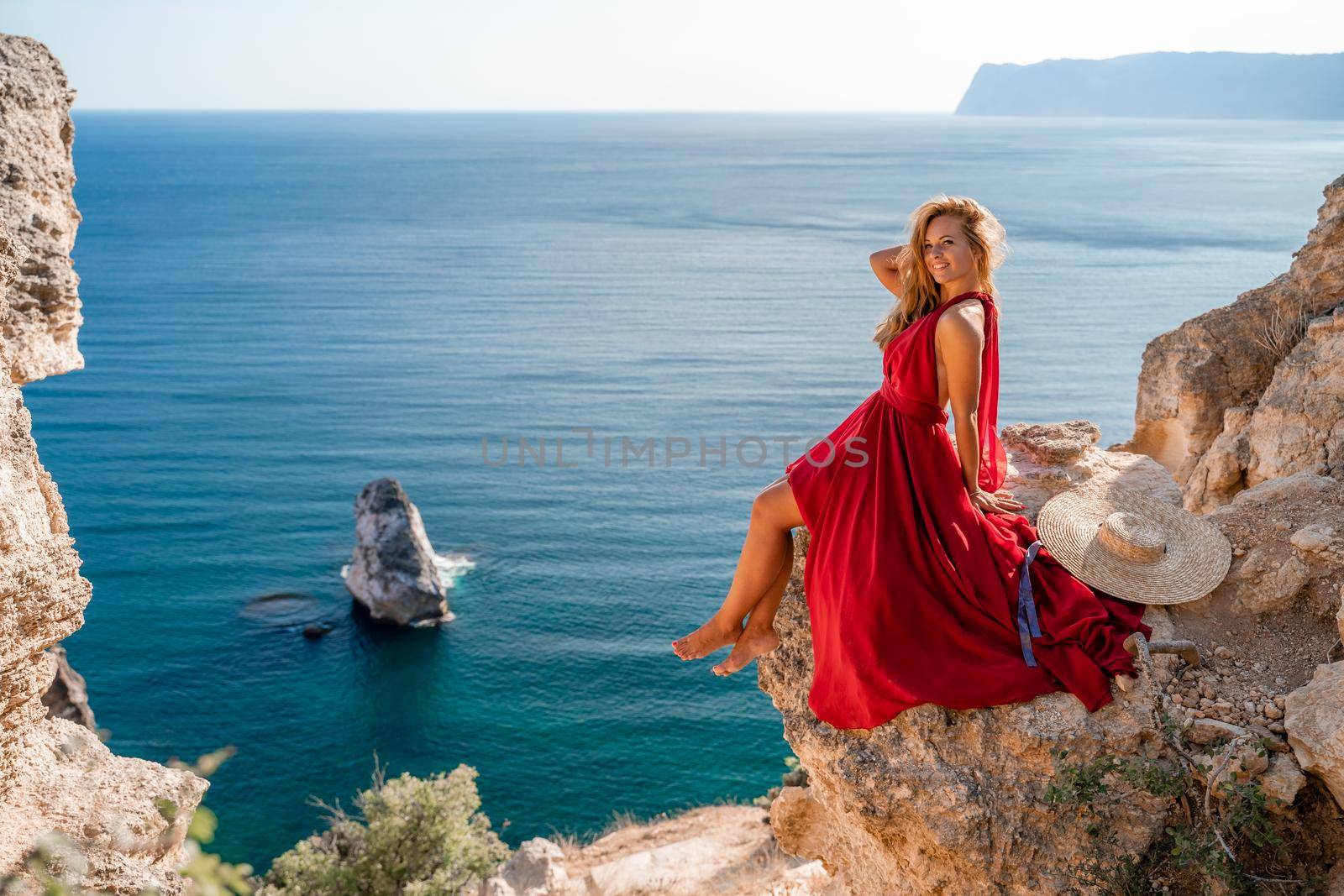 A girl with flowing hair in a long red dress sits on a rock above the sea. The stone can be seen in the sea. by Matiunina