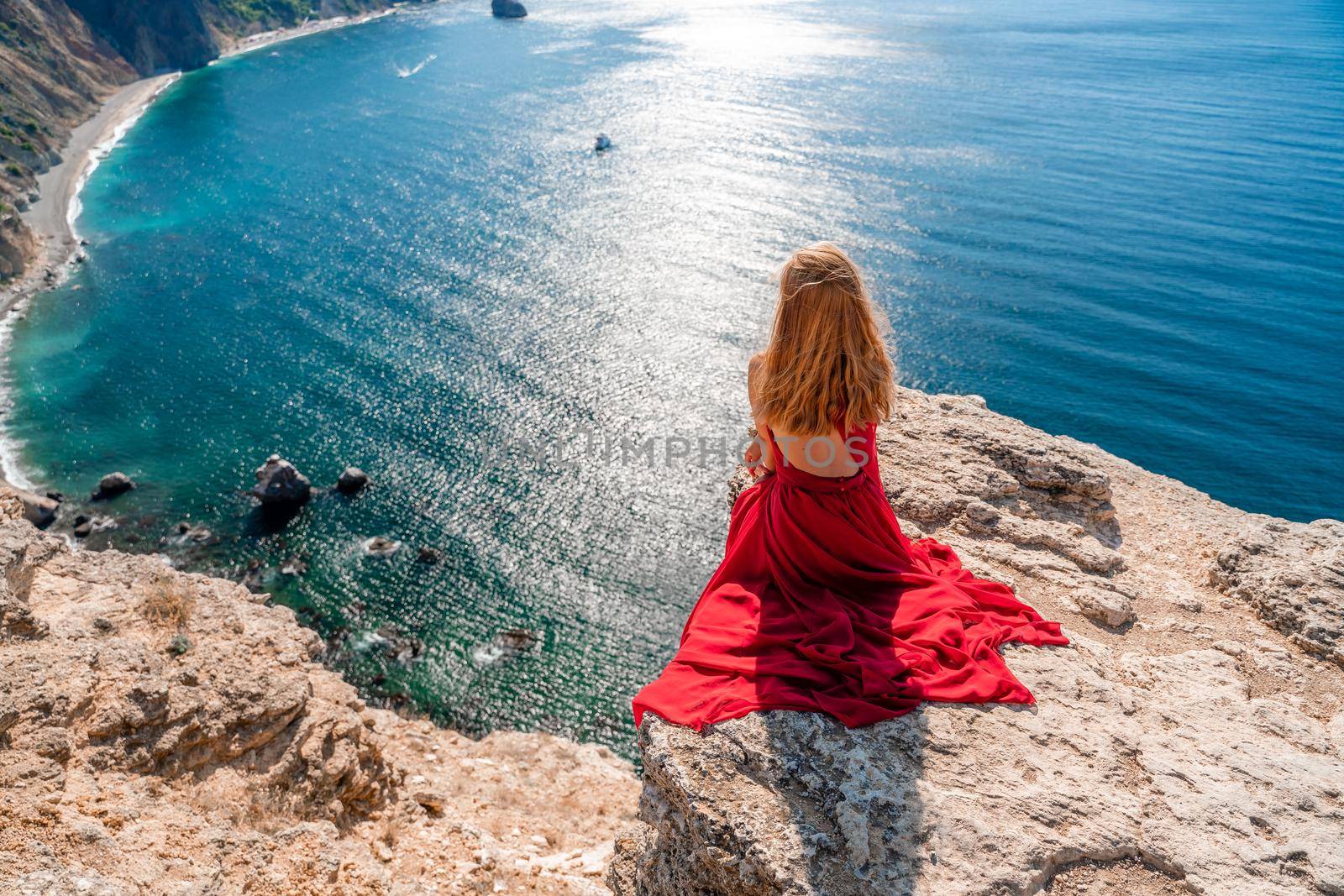 A girl with flowing hair in a long red dress sits on a rock above the sea. The stone can be seen in the sea. by Matiunina