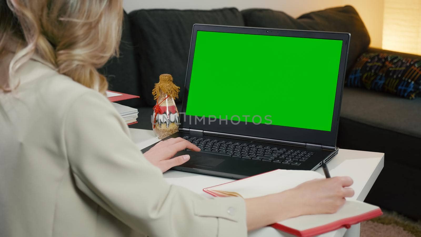 Chromakey Mockup. Woman writing and using laptop with blank green screen. Home office work. Makes notes from laptop sitting on table. Blonde lady