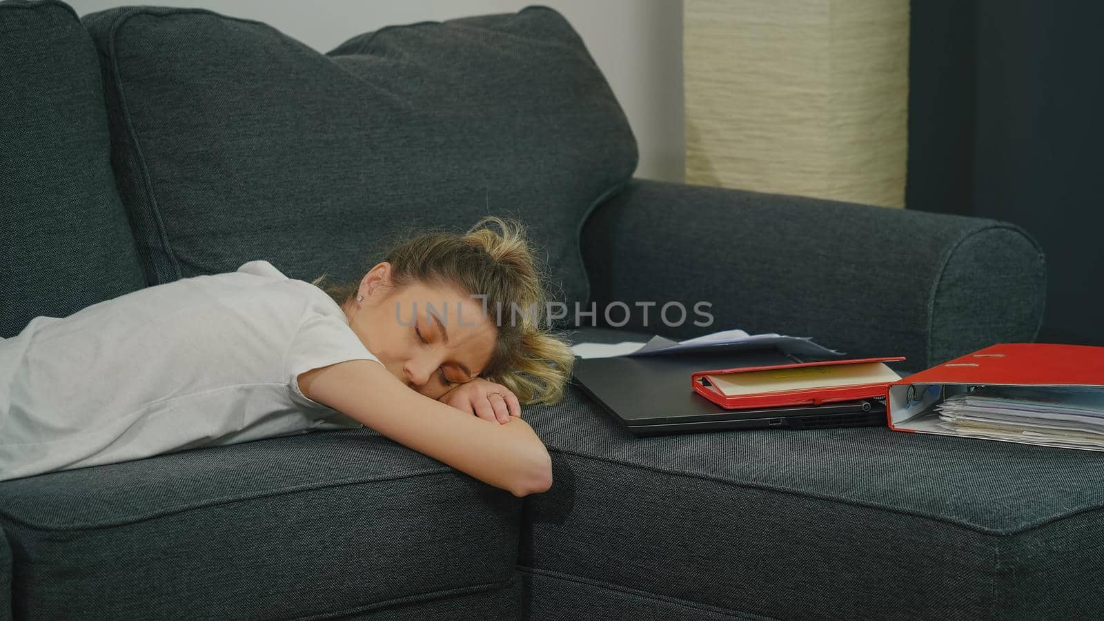 The young woman slept on the couch next to laptop and office papers after a hard day's work. Overworked work from home. First job after college and and many new things to learn. First work experience.