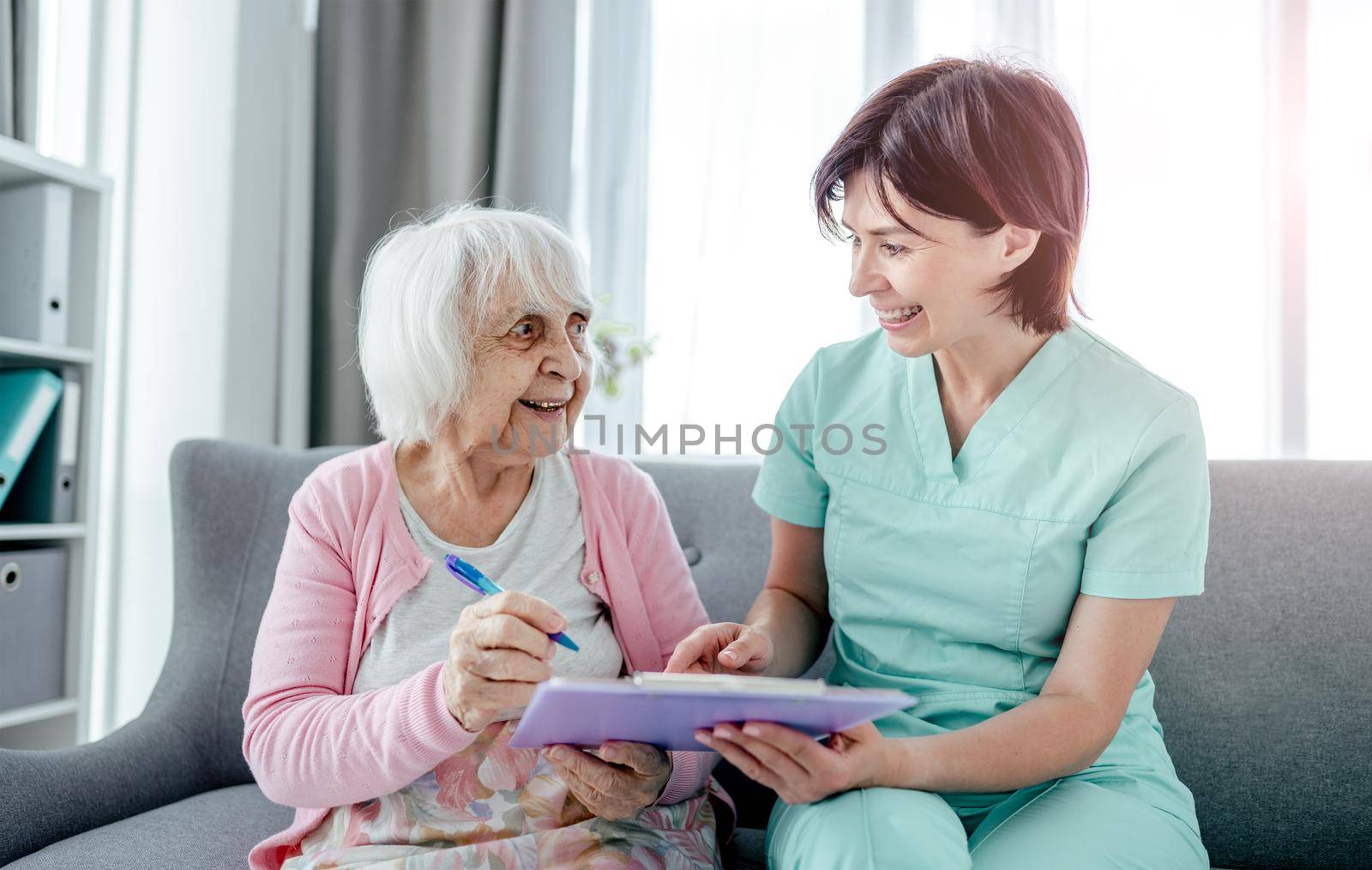 Elderly woman and nurse sign documents at home. Healthcare worker girl cares about senior female person indoors