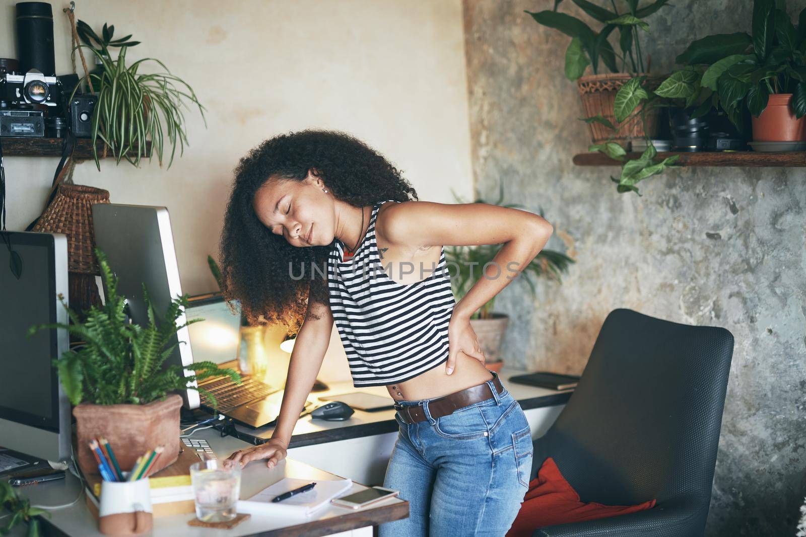 Shot of a young african woman sitting alone and suffering from back ache while using her computer to work from home stock photo