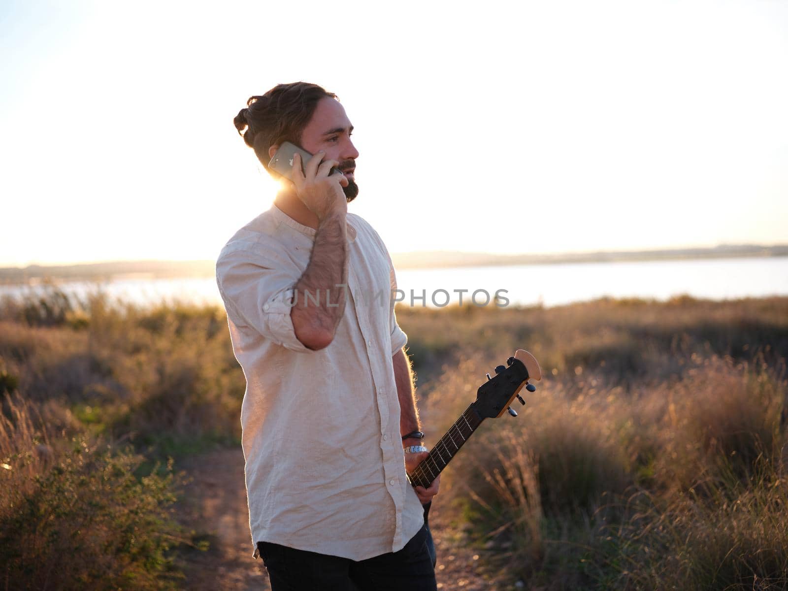 male guitarist calling on the phone in the nature blocking out the sun with his body, horizontal waist up view