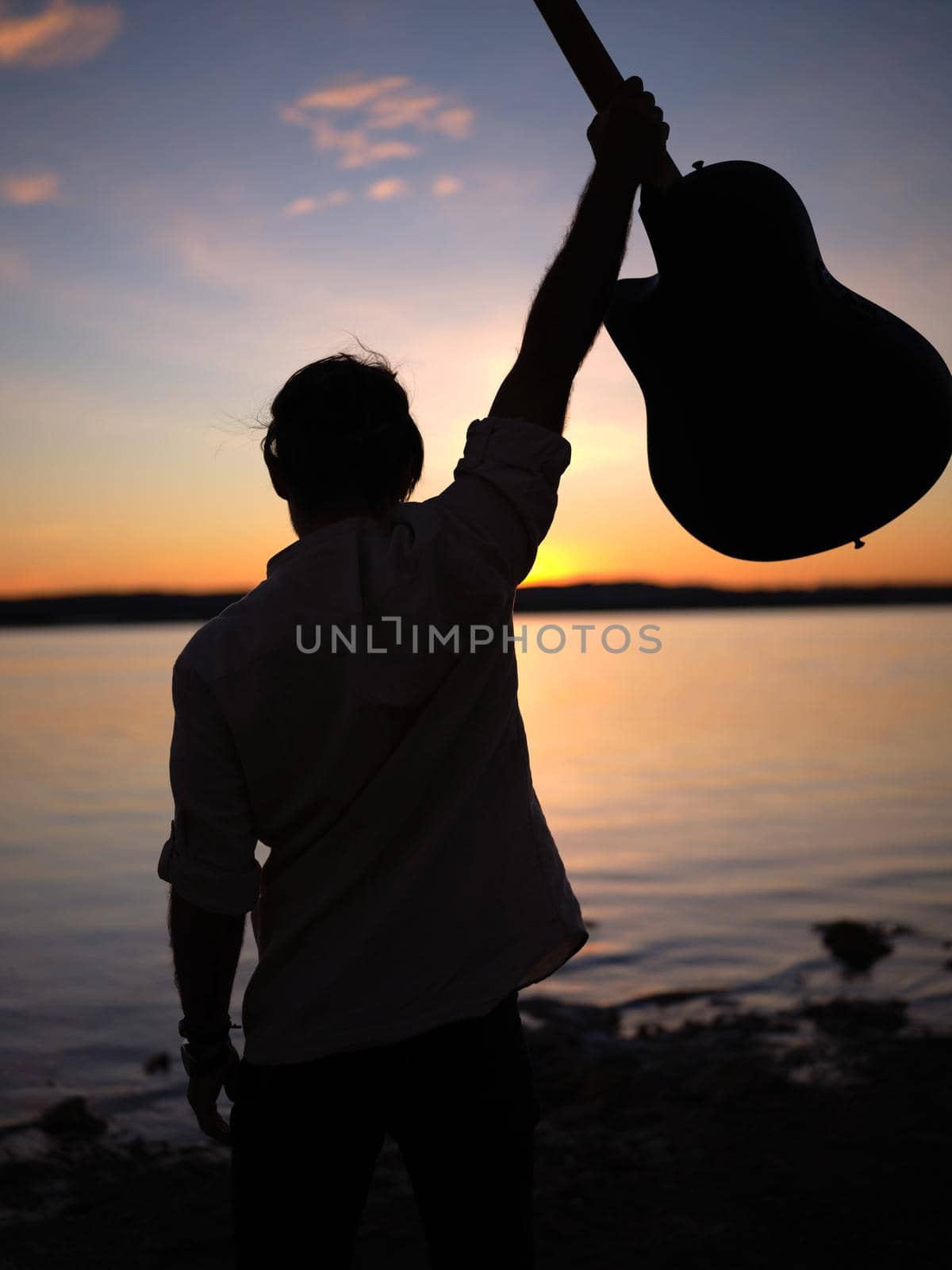 man raising his guitar on a lakeshore at nightfall by WesternExoticStockers
