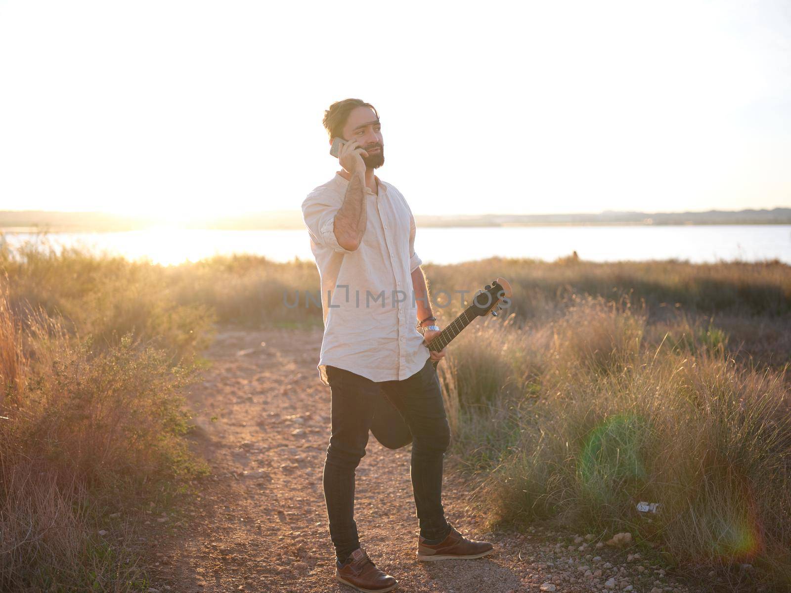 man standing with a guitar in his hand and talking on the phone, sunset on the horizon in the background