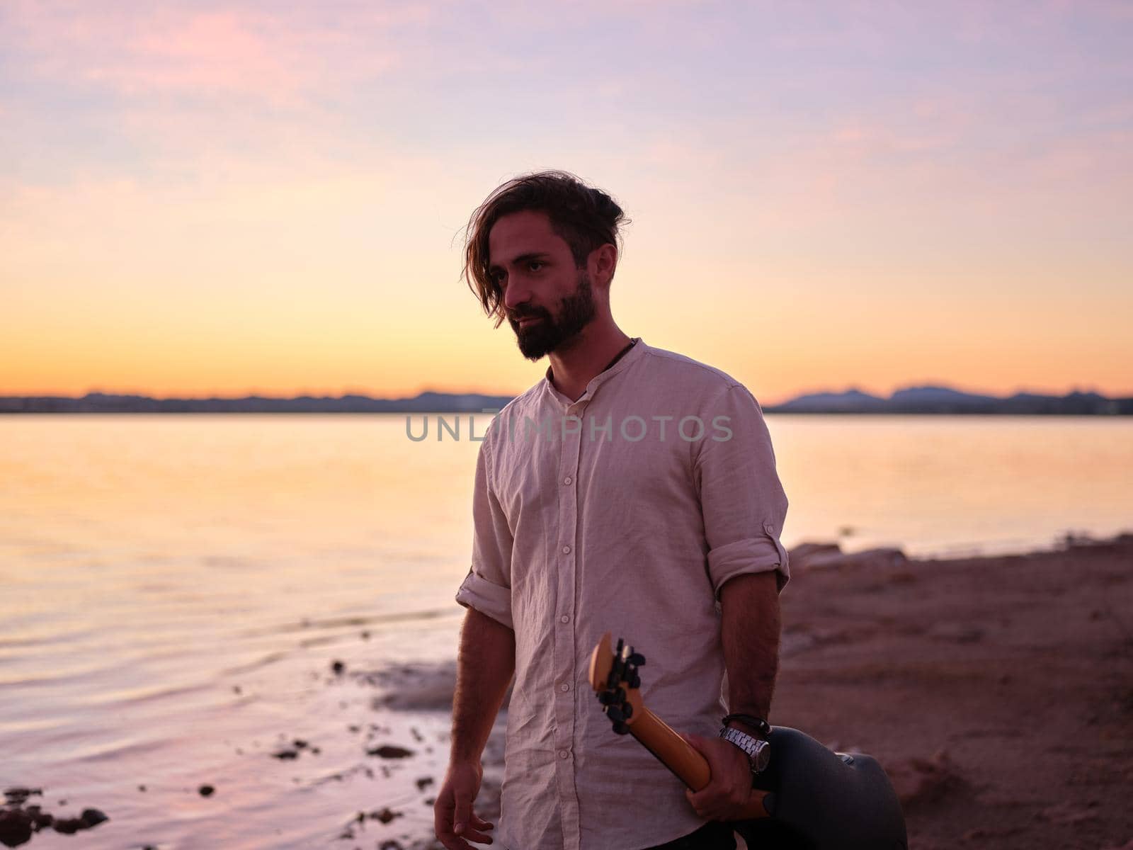 man gazing intently ahead, holding his guitar in one hand, nightfall on the lake, horizontal landscape