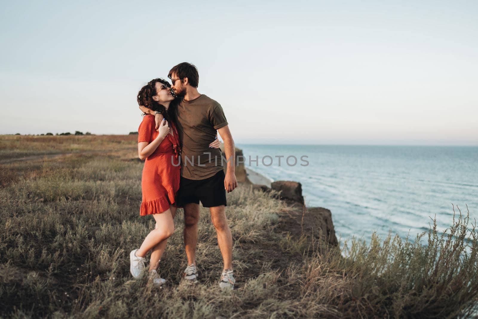 Cheerful Young Couple Standing on Background of Sea, Happy Man Hugging Curly Woman in Red Dress at Sunset