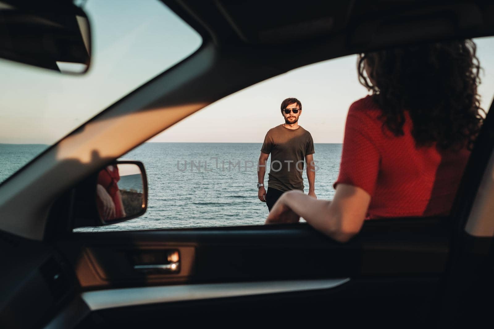 View Through Window of the Car, Man Standing in Front of Curly Woman in Red Dress on Background of Sea at Sunset