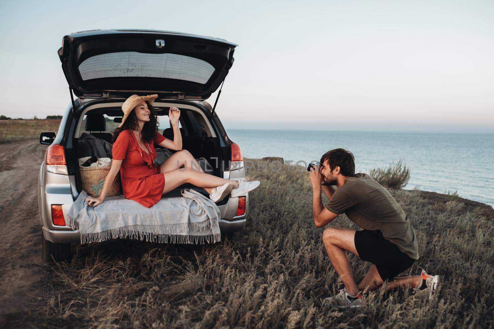 Man Photographing Woman in Red Dress and Hat That Sitting Inside Opened Trunk of Car on the Background of Sea