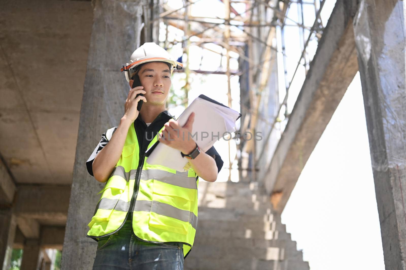 Young engineer man in helmet talking on mobile phone under new building.