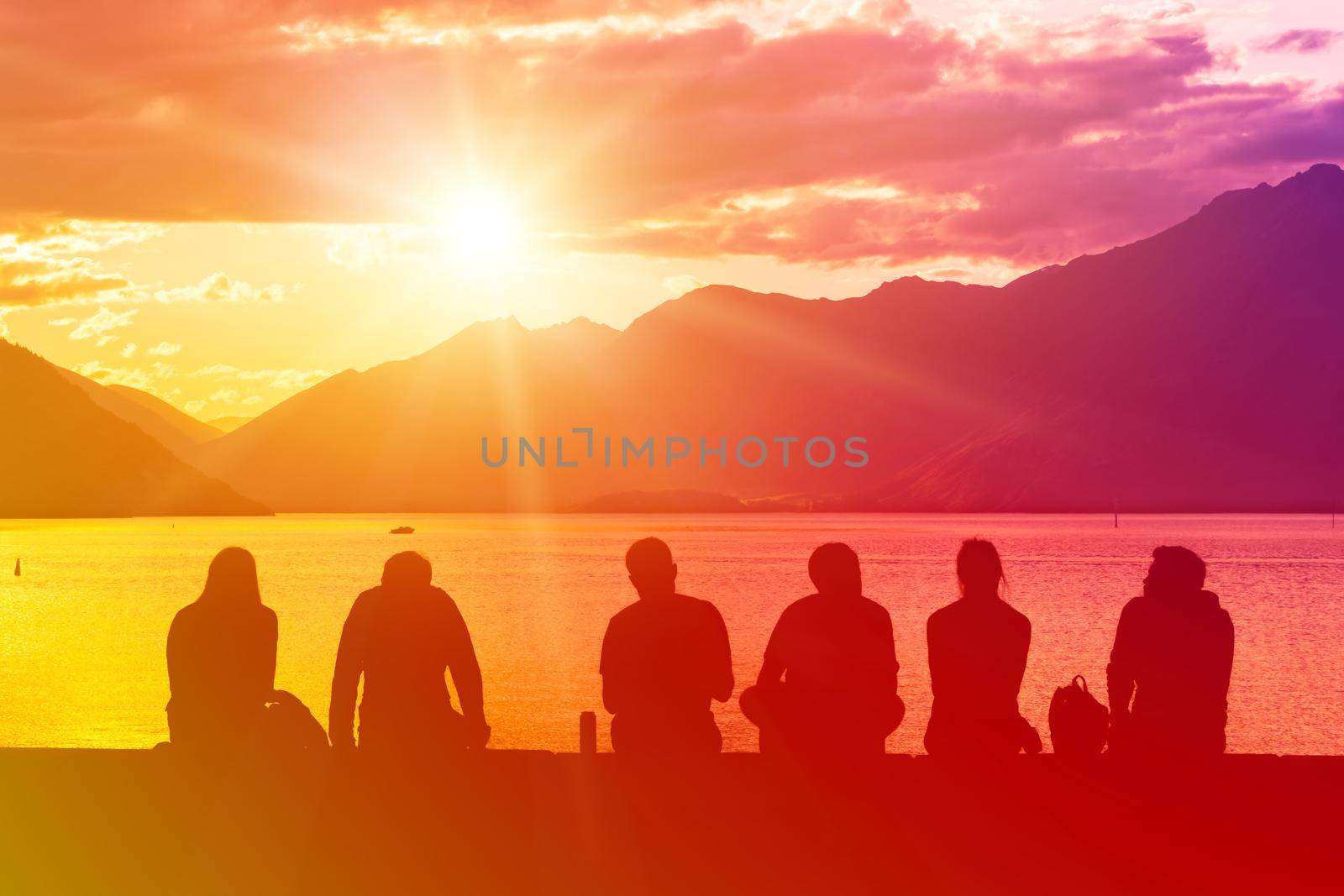 Silhouette group of young people sitting on lake beach against mountain landscape at sunset showing concept of youth friendship, togetherness, success and teamwork of teenager friends in university.