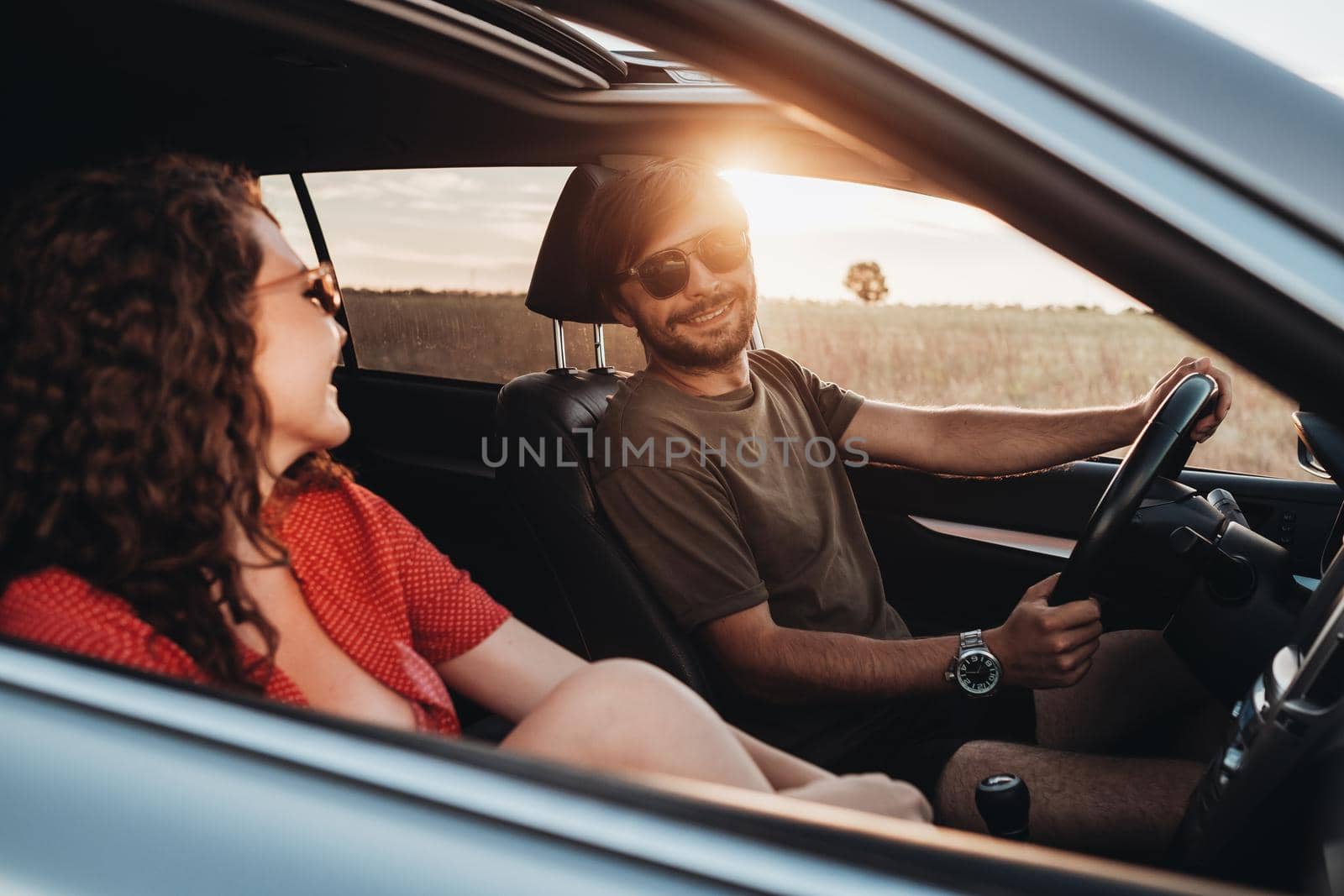 Cheerful Man and Curly Brunette Woman Sitting in Car, Young Couple Enjoying Road Trip at Sunset