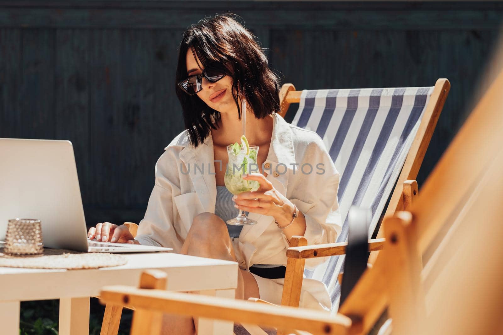 Young Brunette Woman in Sunglasses Working on Laptop and Drinking Cocktail While Sitting on Terrace of the Restaurant, Freelancer at Work